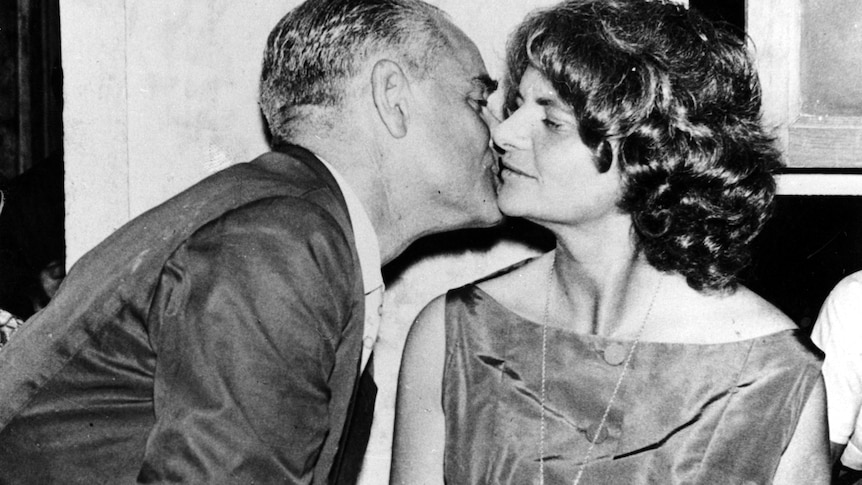 A B&W photo of a man leaning into kiss the check of a woman with a curly bob. Her face is in front and her eyes are closed