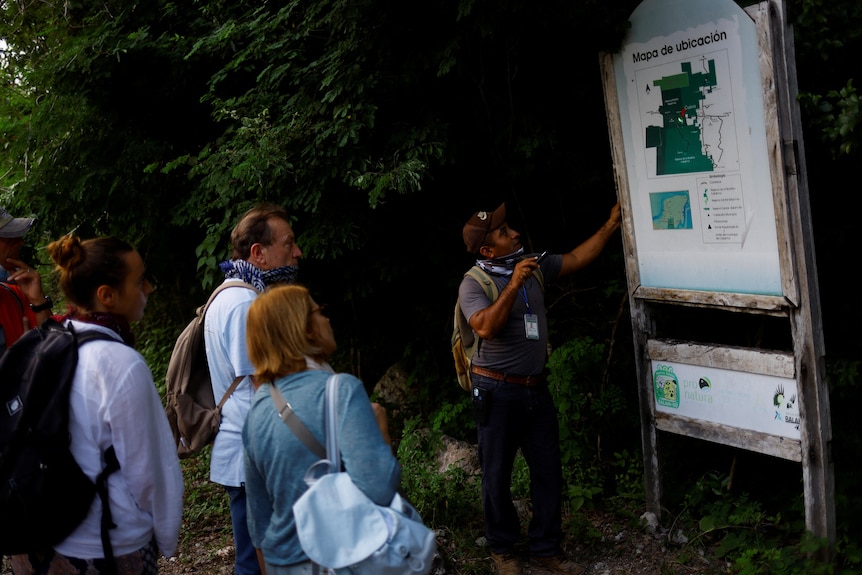 A tour guide stands in front of a map sign as three people watch. 