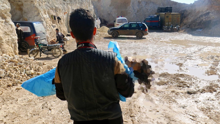 A man holds a child's body wrapped in blue sheeting. The child's face is blurred.