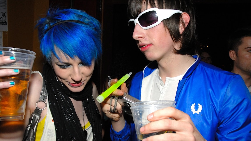 Two 2007 ravers with blue and black hair in blue jackets, black glitter scarves hold beers, glowsticks and a digital camera