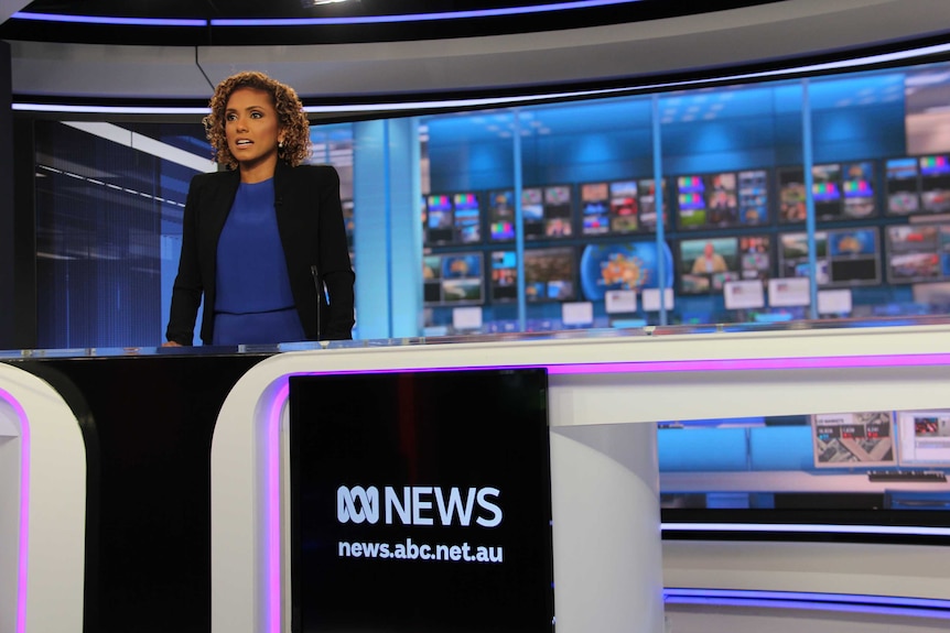 Carvalho standing at news desk in studio with huge bank of TV monitors in background.