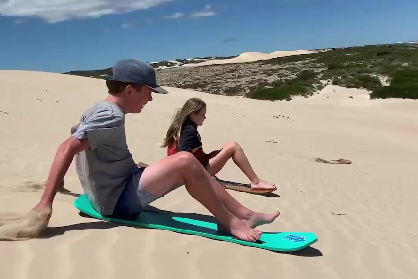 Boy and girl sitting on sandboards going down a sandune with greenery and sandhills in distant background.