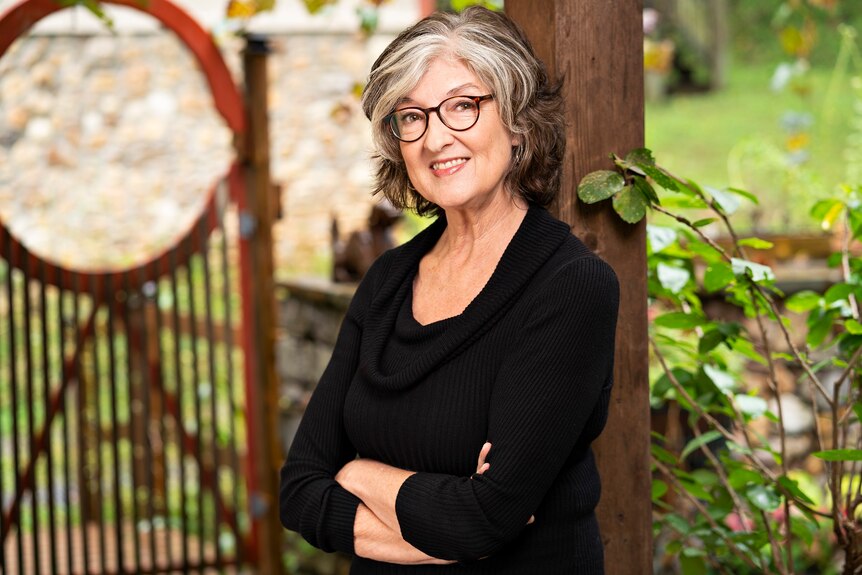 A woman with grey hair and glasses leans against a wooden post with greenery in the background