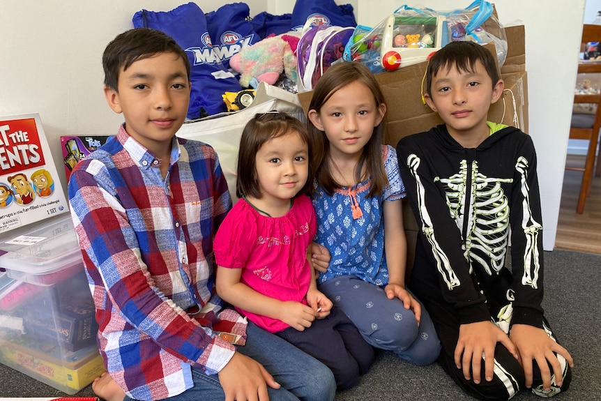 Four young children sit side by side with boxes of toys behind them.