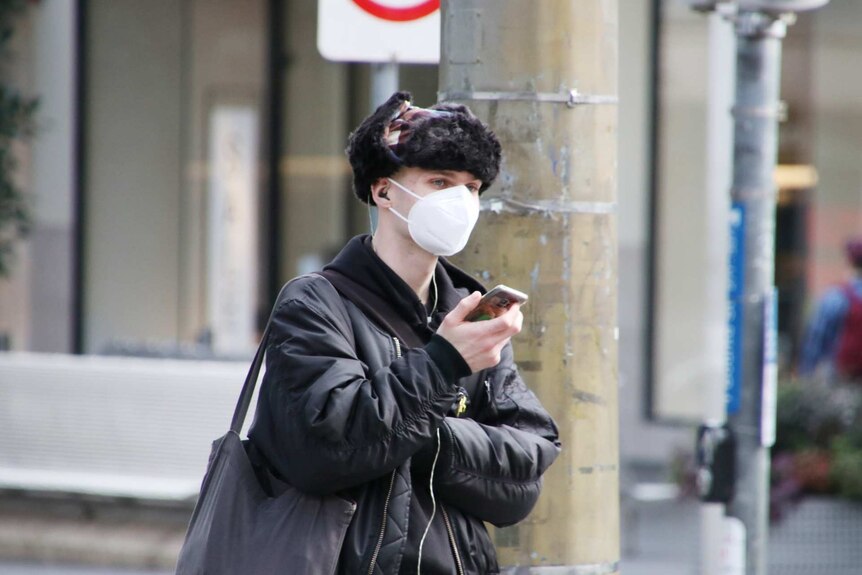 A man in a fur hat, wearing a mask, checks his phone in the city