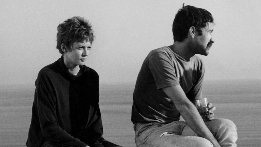A short-haired young woman and a young man with a mustache sit atop a wall with the ocean in the background