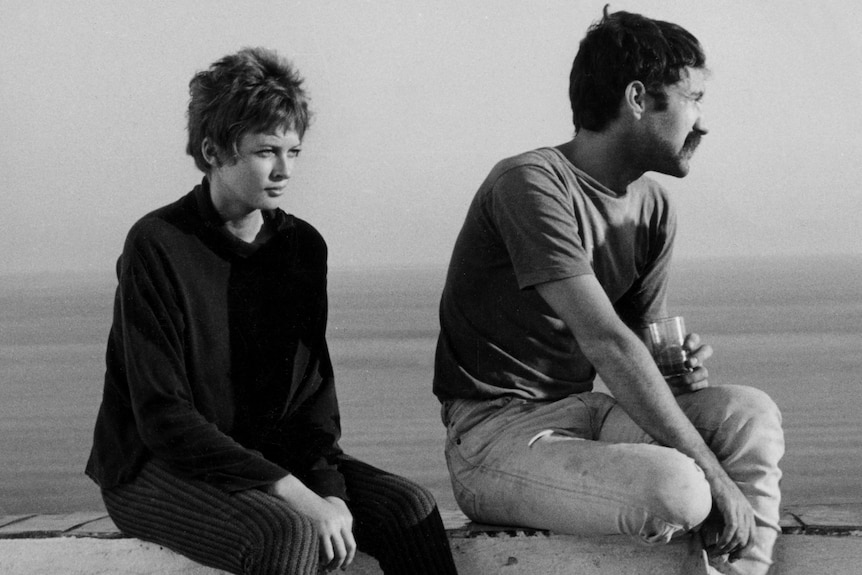 A short-haired young woman and a young man with a mustache sit atop a wall with the ocean in the background