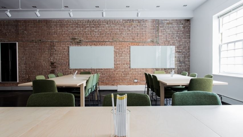Photo of a class room with a red brick wall, desks, green chairs and a jar of white pencils 