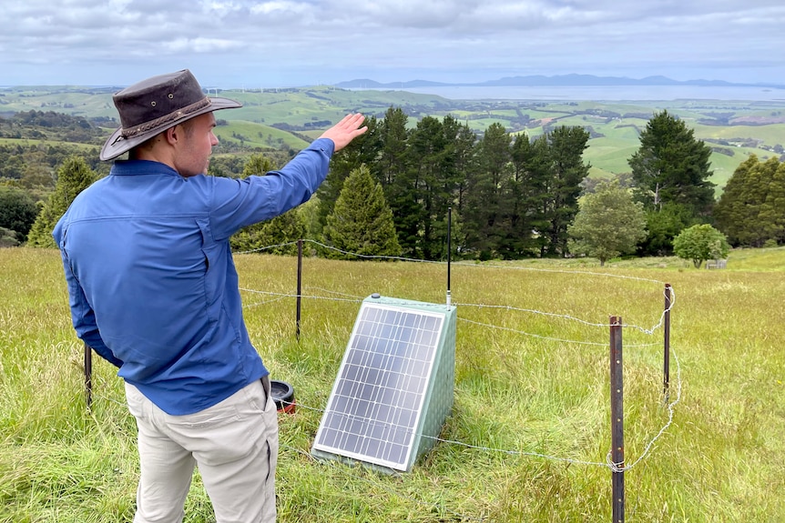 A man stands by a seismometer, featuring a solar panel mounted on a green metal hut, pointing off into the distance.