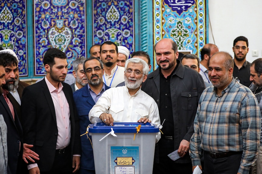 An older Iranian man with a white beard casts a ballot in front of a group of men.