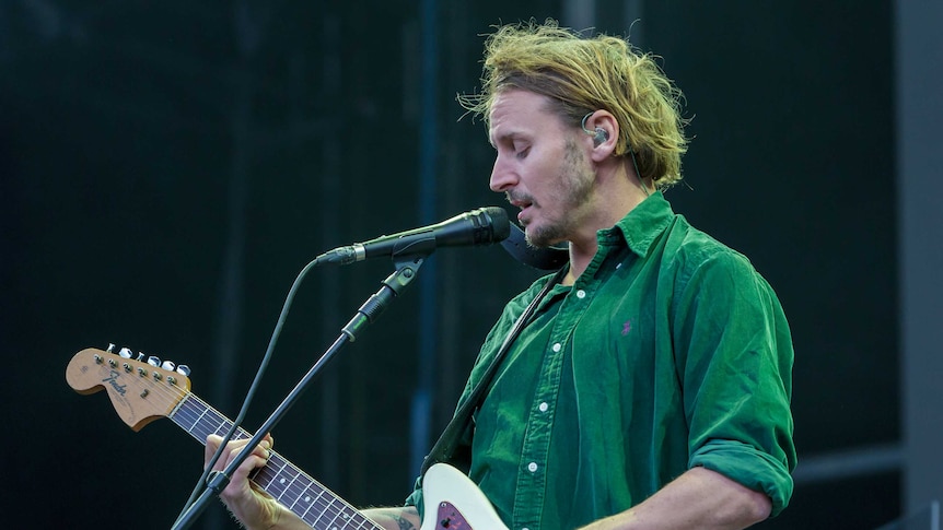 Ben Howard sings into a microphone while playing guitar.