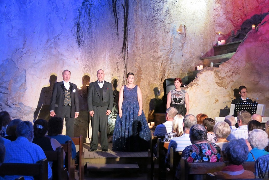 Four singers perform in front of seated guests. Behind them, limestone walls with shades of blue and pink from lights