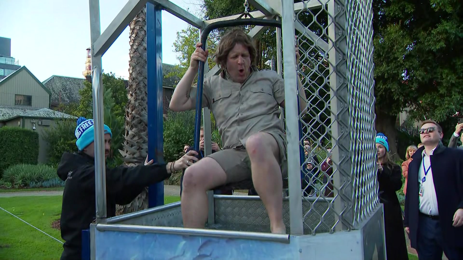 A man in a blonde wig and khaki shirt and shorts winces while sitting on a raised seat inside a steel cage.