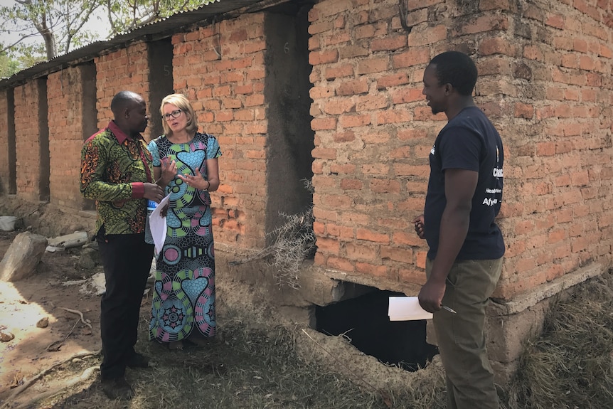 A western woman speaking with an African man near a brick building.