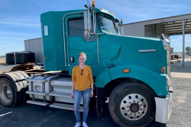 A slim woman with her hair in a bun, standing in front of a truck, smiling.