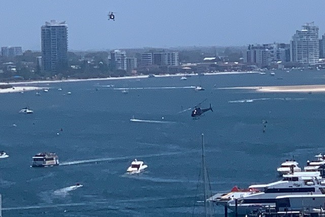 Two helicopters in the sky above a busy section of ocean in front of a city.
