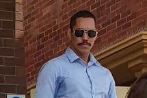 Man in sunglasses looks at camera outside court, moustache, close cropped hair.