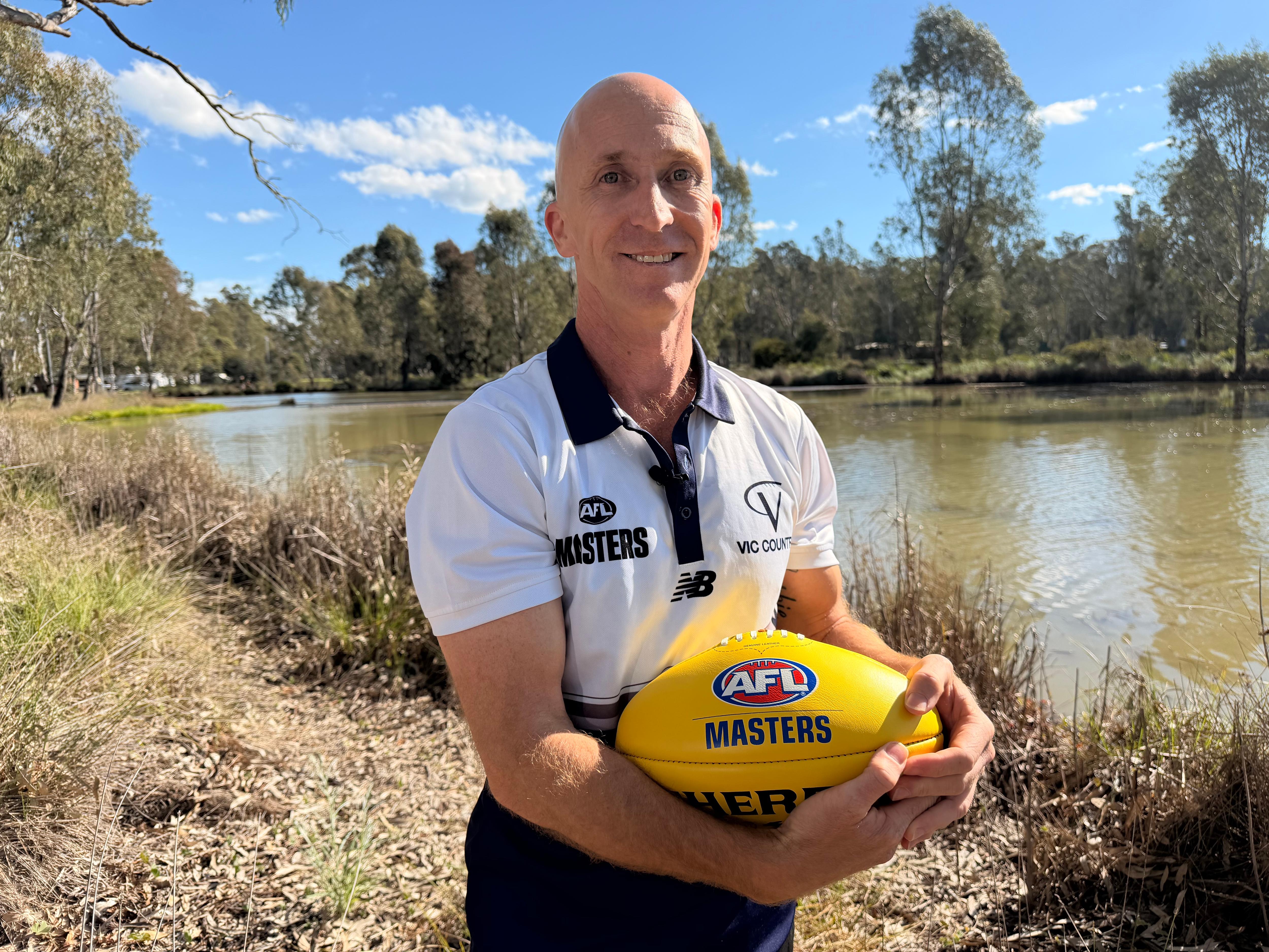Victoria Country AFL Masters president Jason Aspland standing in front of a river, holding a yellow football