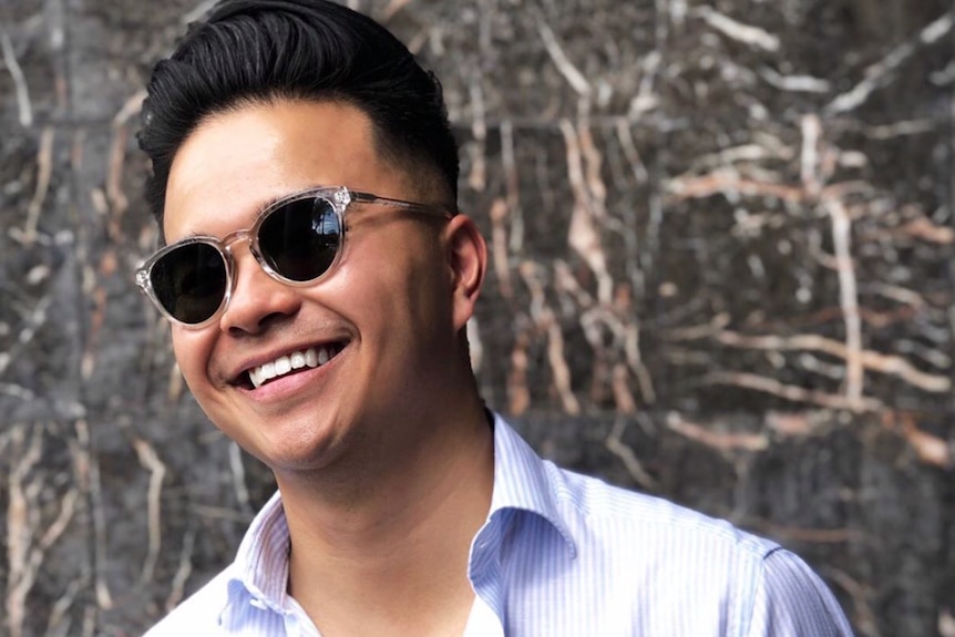 A young man wearing dark sunglasses is smiling standing in front of a grey wall. He's wearing a light coloured shirt.