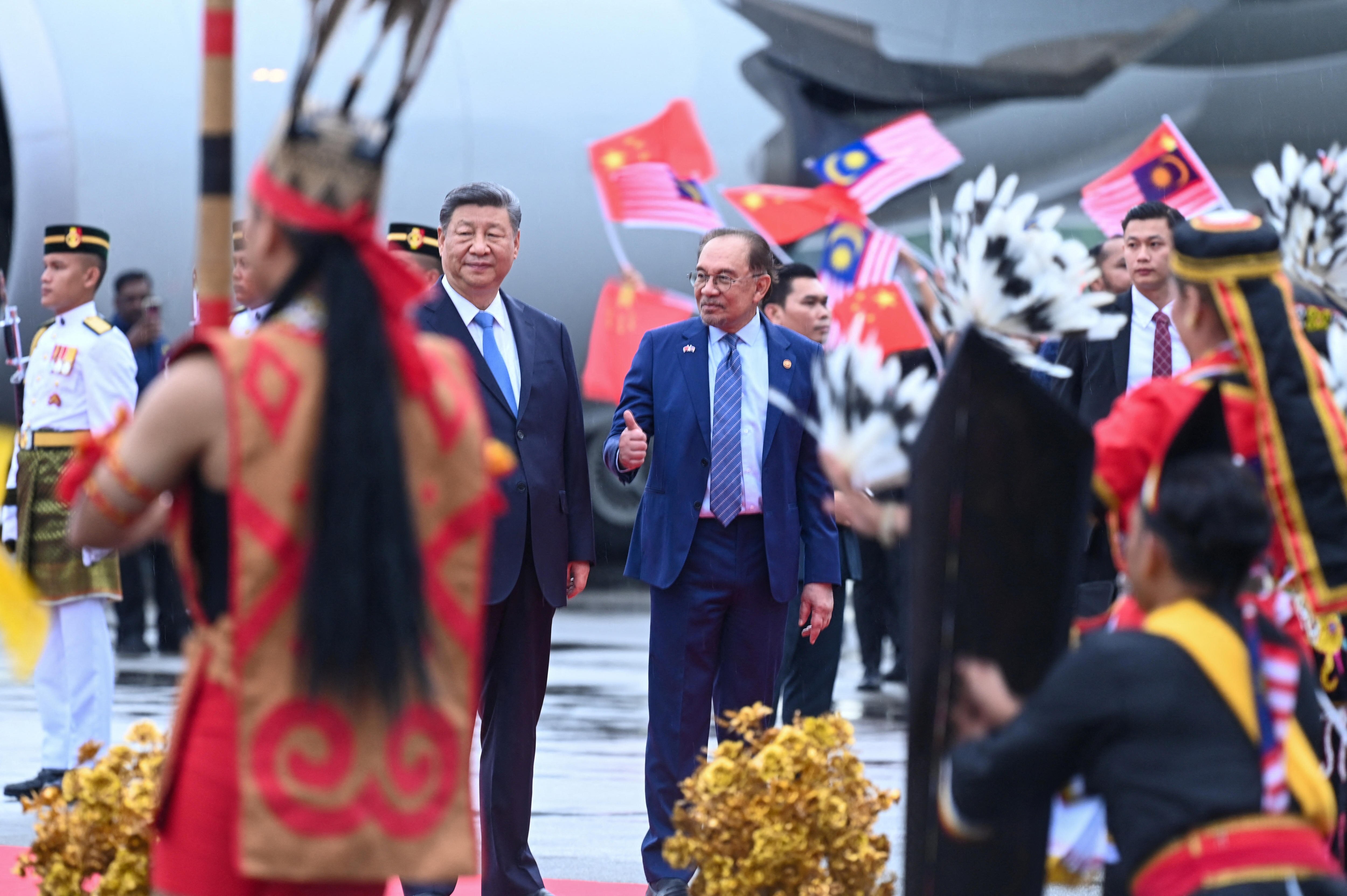 两个穿着西装的男人站在停机坪上，看着戴着羽毛帽子的舞者，而其他人则放弃旗帜