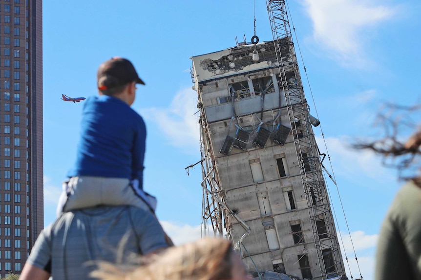 A small boy sits on a man's shoulders and watches a building in the distance get hit by a wrecking ball.