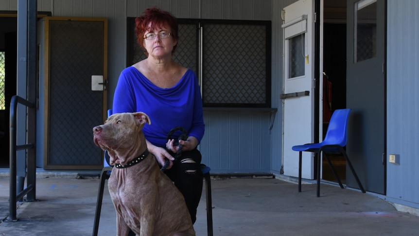 A woman in a bright blue top sits on a chair. At her feet is her grey pet dog.