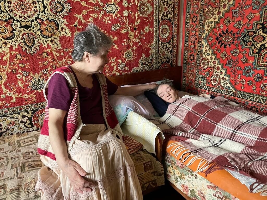 A woman with cropped grey hair sits next to a woman lying in a bed covered in blankets 