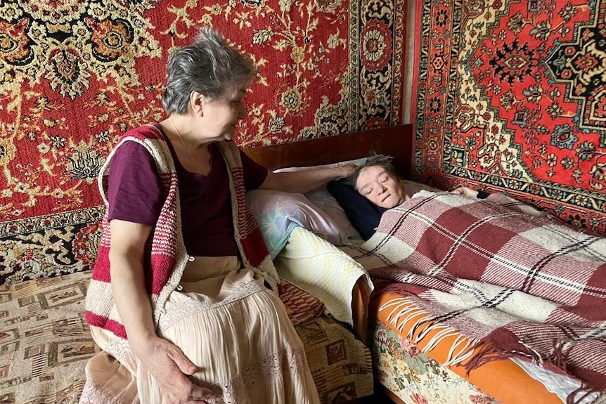A woman with cropped grey hair sits next to a woman lying in a bed covered in blankets 