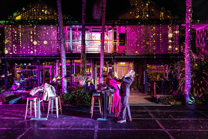 A man and woman dramatically embrace each other on an outdoor stage. A building is bathed in purple light behind them.