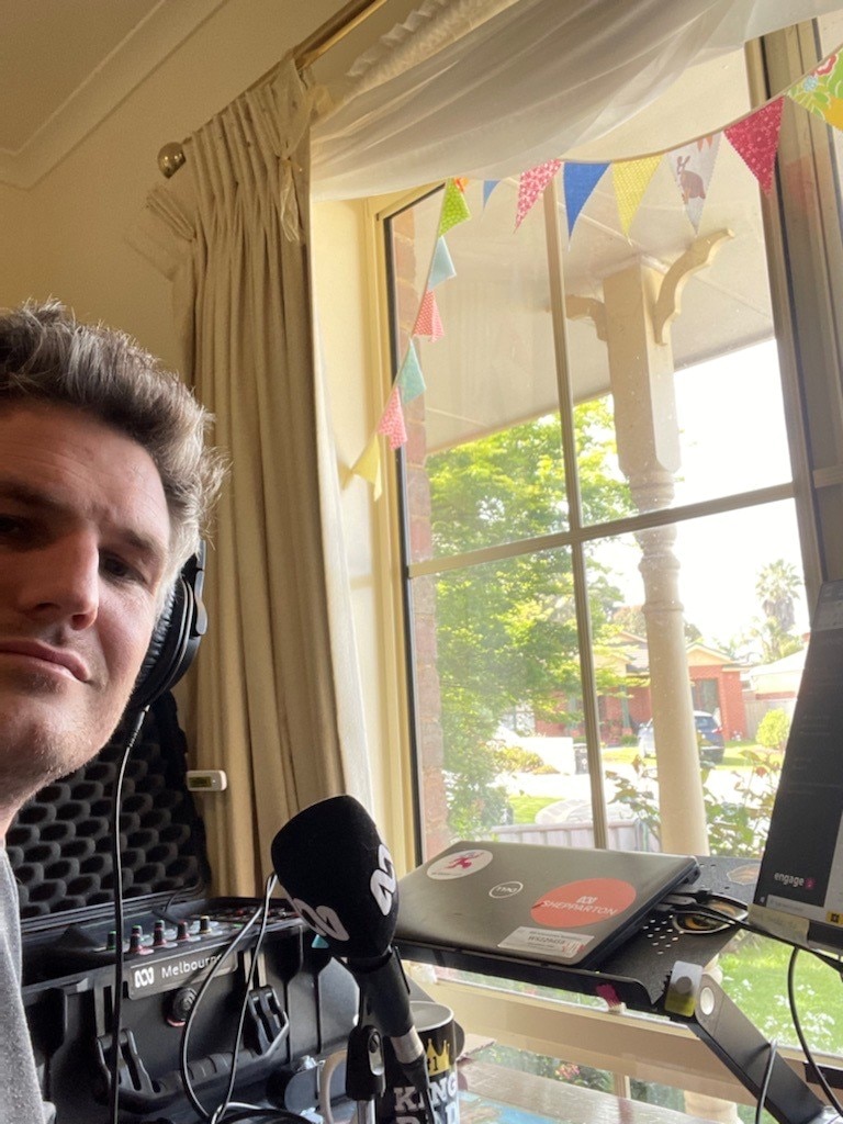 Man sitting at desk at lounge room window with headphones on and microphone and laptop in front of him.