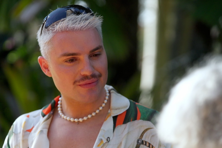A blonde man with a dark moustache and sunglasses on his head looks down and to the right. He wears a pearl necklace