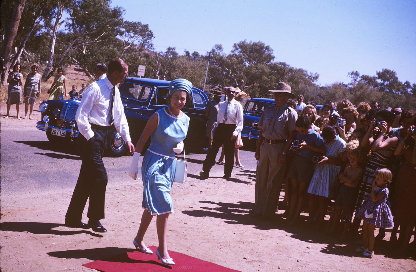The Queen and the Duke of Edinburgh walk from the red dirt onto the red carpet as people line up to see them.