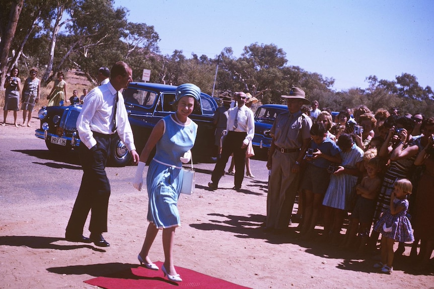 The Queen and the Duke of Edinburgh walk from the red dirt onto the red carpet as people line up to see them.