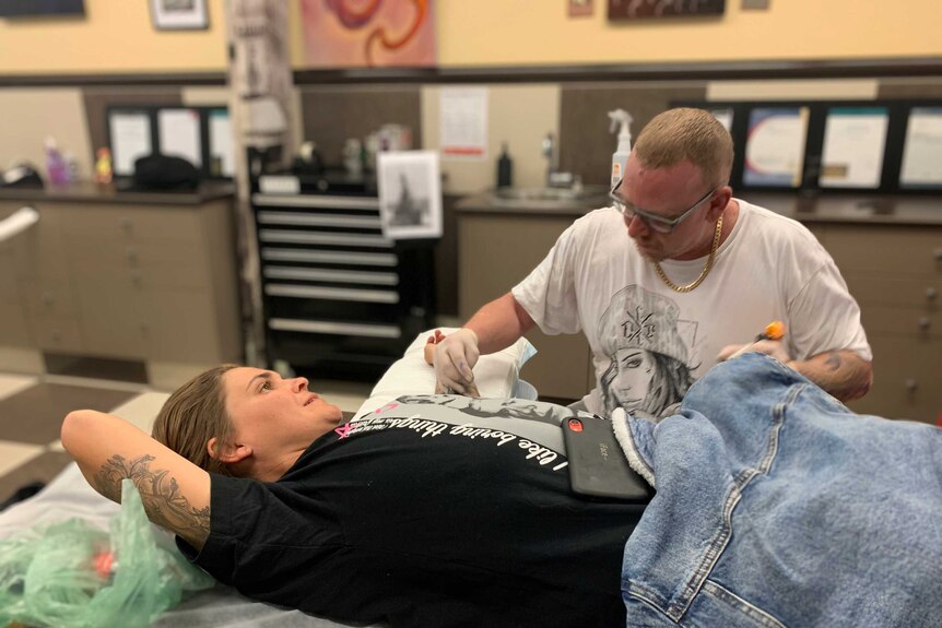 Customer Skye Ronan lying down as tattoo artist inks her left arm with Chinese good luck coin symbol