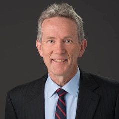 A headshot of an older man with neat grey hair, wearing a dark suit.