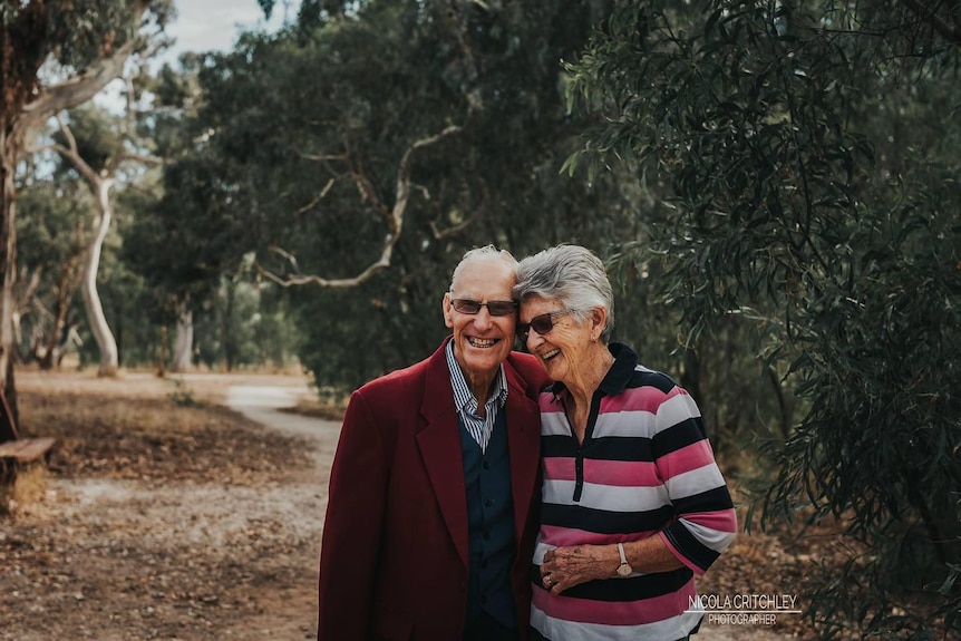 An older couple smiling, leaning into each other, with a bush track behind them