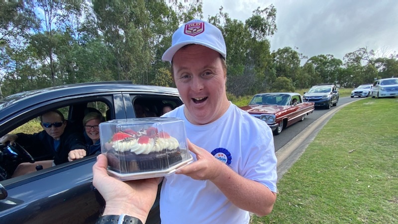 arm taking cake from man smiling at camera, car in the background and a line up of cars.