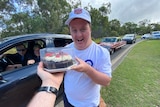 arm taking cake from man smiling at camera, car in the background and a line up of cars.