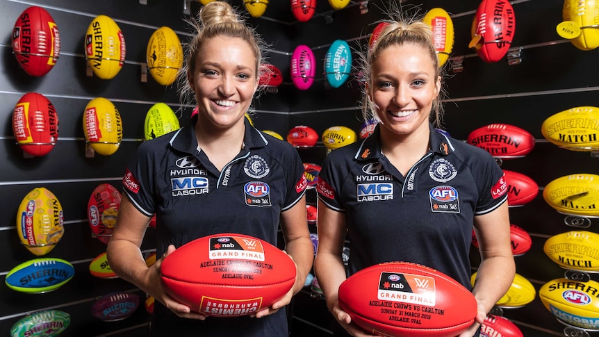 Two identical twin sisters hold footballs in their Carlton gear ahead in 2019 AFLW grand final week.