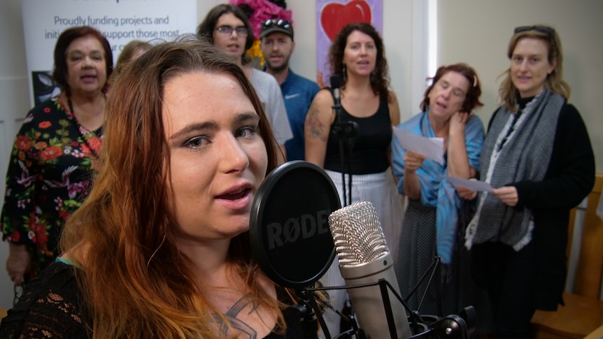 A woman with long brown hair standing at a recording microphone, with a group of people behind her.