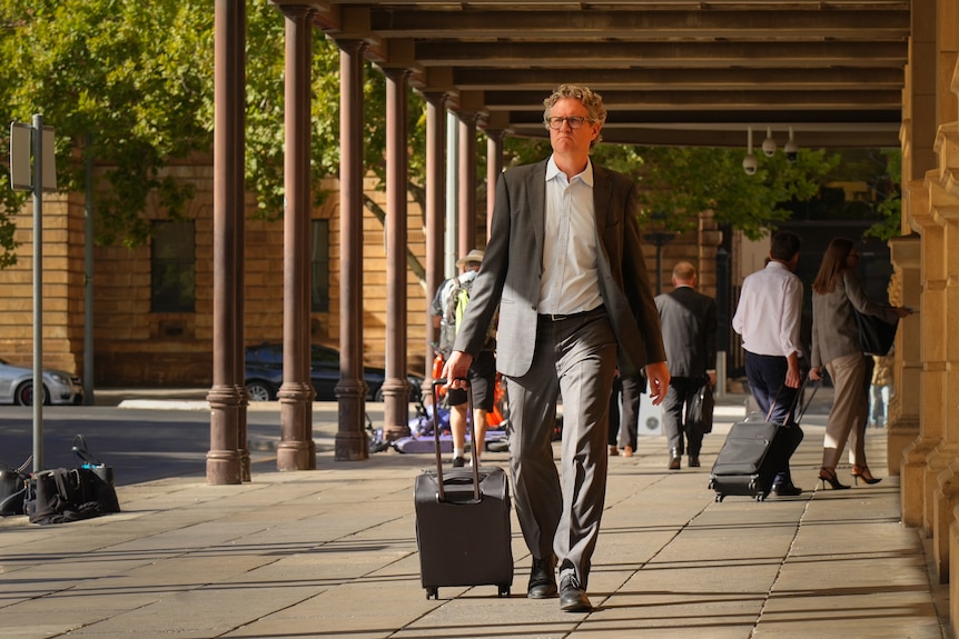 A man in grey suit and white shirt walks past Samuel Way Building with suitcase.