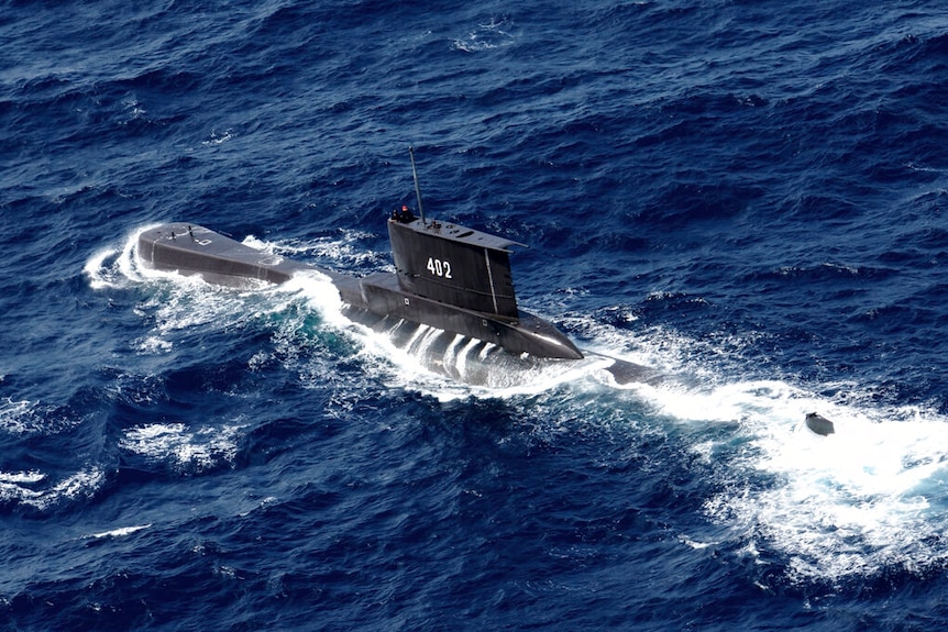 Una foto aérea muestra un submarino rompiendo la línea de flotación en un día claro en aguas de un azul profundo.