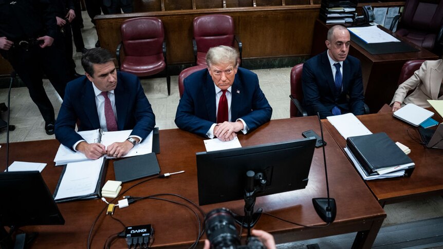 Donald Trump between two men at desks in a courtroom looking sullen