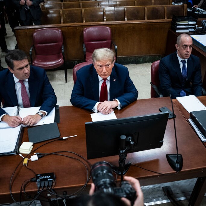 Donald Trump between two men at desks in a courtroom looking sullen