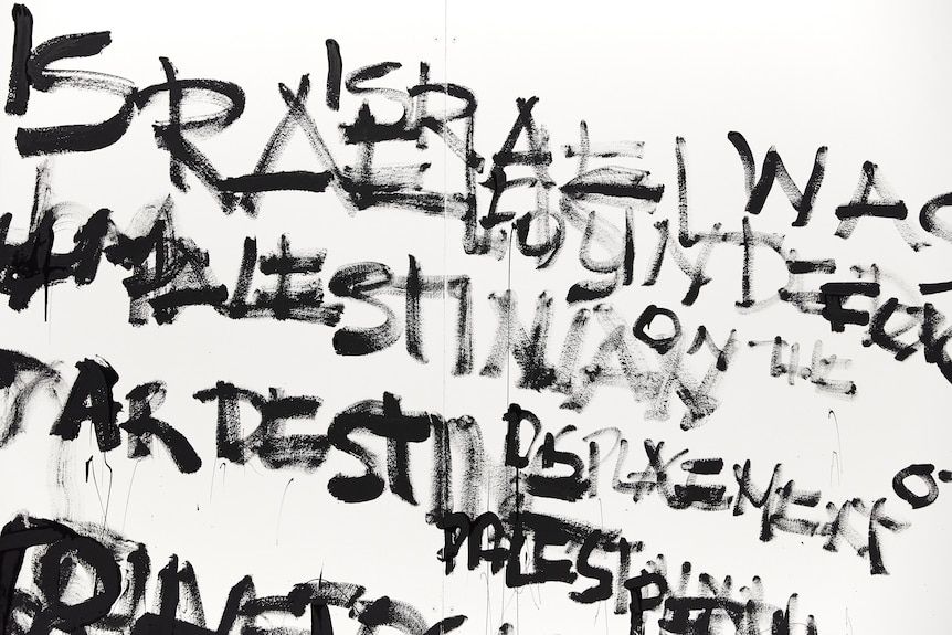 A close up of text painted illegibly in black on a white gallery wall. The words Israel and Palestine are visible.