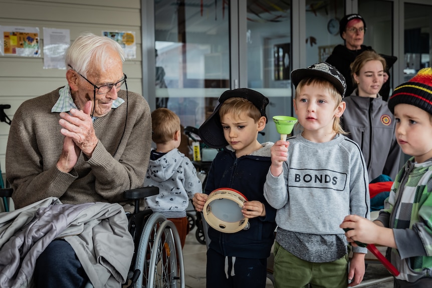 An elderly man sits in a wheelchair surrounded by young toddlers.