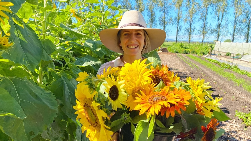 A woman smiles at the camera while holding freshly farmed sunflowers on her vegetable farm