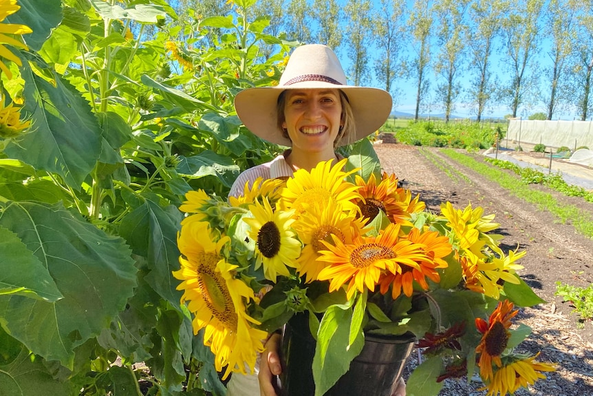 A woman smiles at the camera while holding freshly farmed sunflowers on her vegetable farm