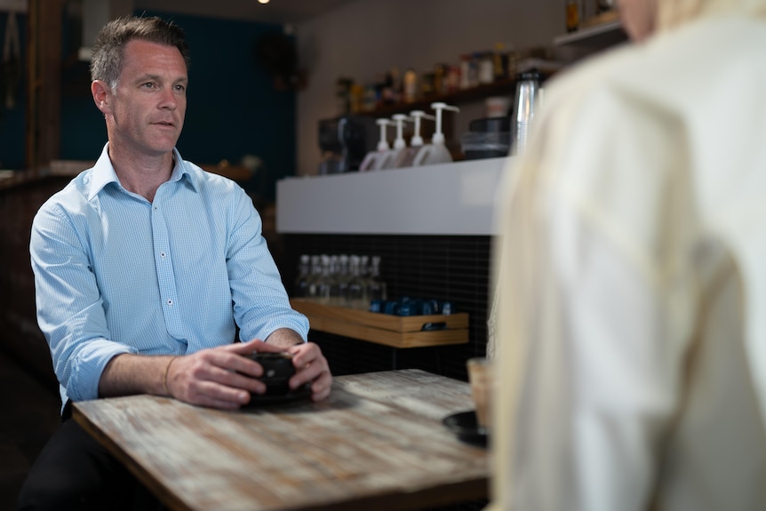 a man sitting down at a cafe holding onto a cup of coffee being interviewed by a reporter