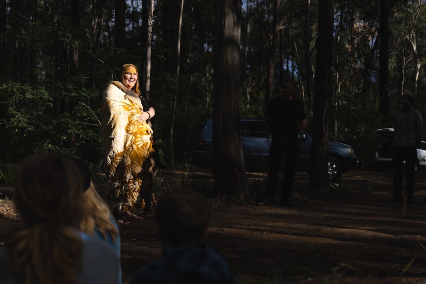 An Aboriginal woman stands in a bush area, in a long robe, smiling.